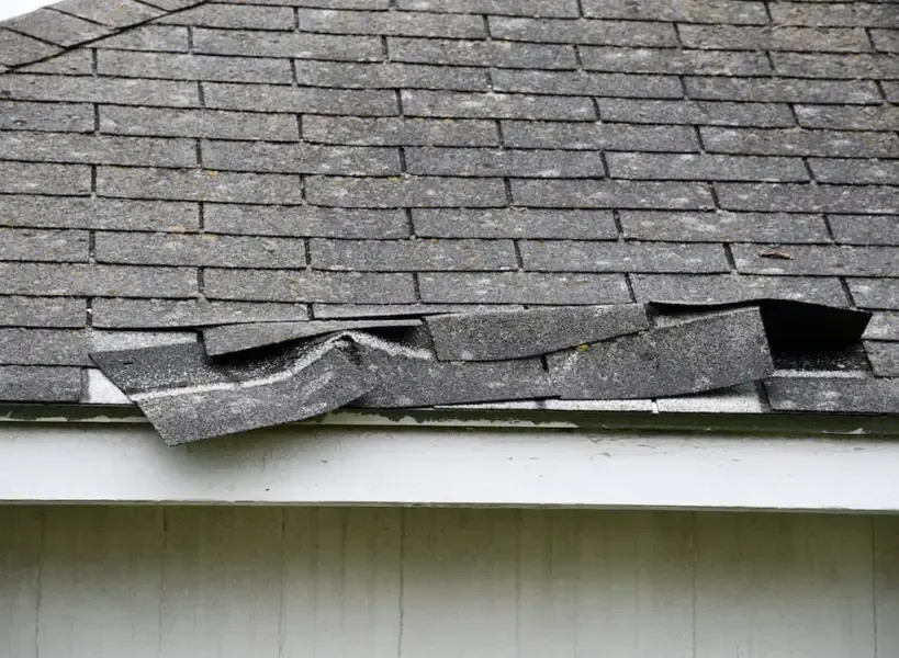 Wind-lifted and torn asphalt shingles along a roof edge, the opening behind many roof leaks